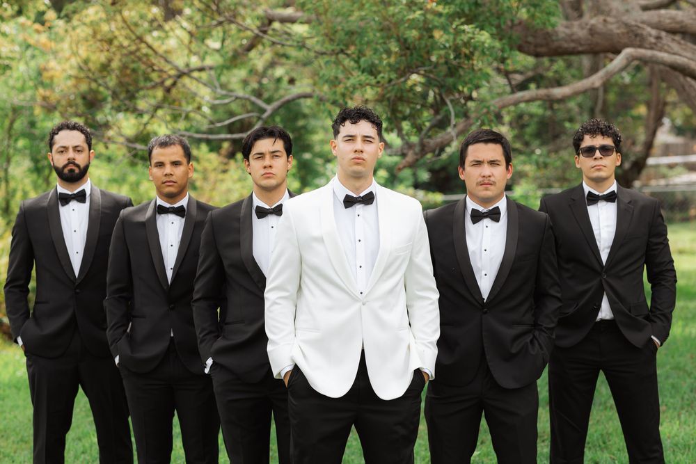 A groom in a white dinner jacket stands centered among groomsmen wearing classic black tuxedos in an outdoor garden setting.