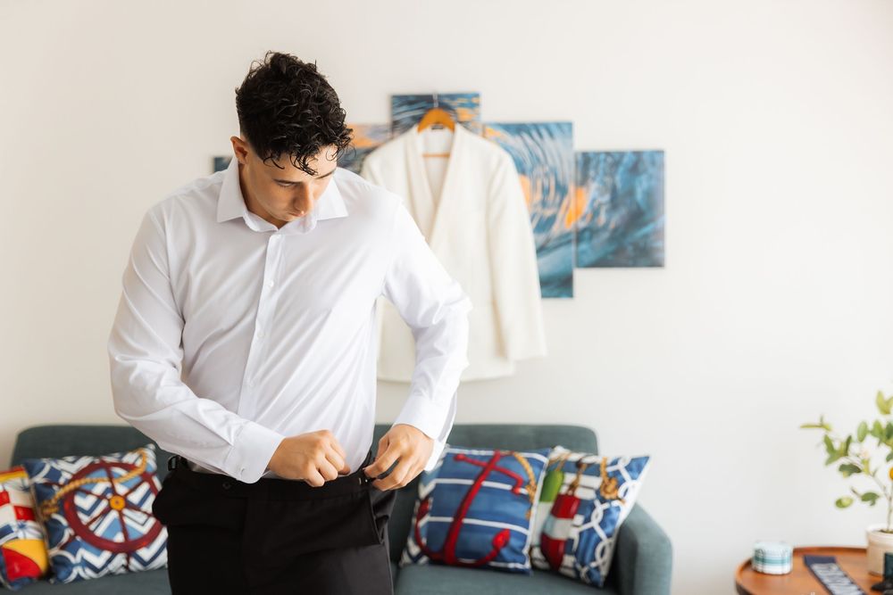 Someone buttons up a crisp white dress shirt while standing in a bedroom with colorful throw pillows and artwork on the wall.