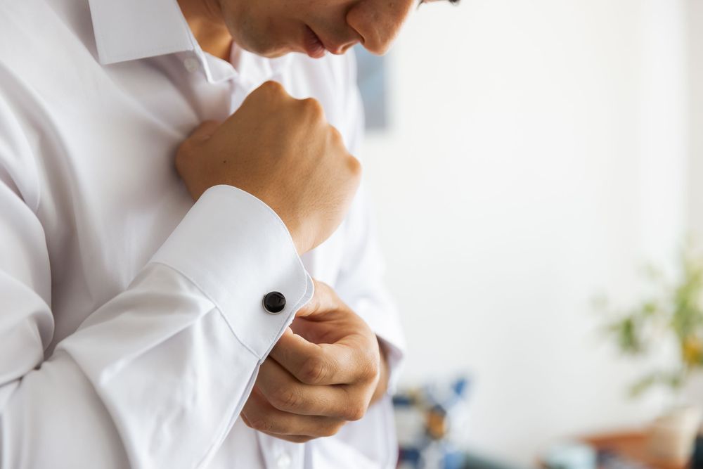 Close-up view of hands adjusting a white dress shirt cuff with cufflinks against a bright background.