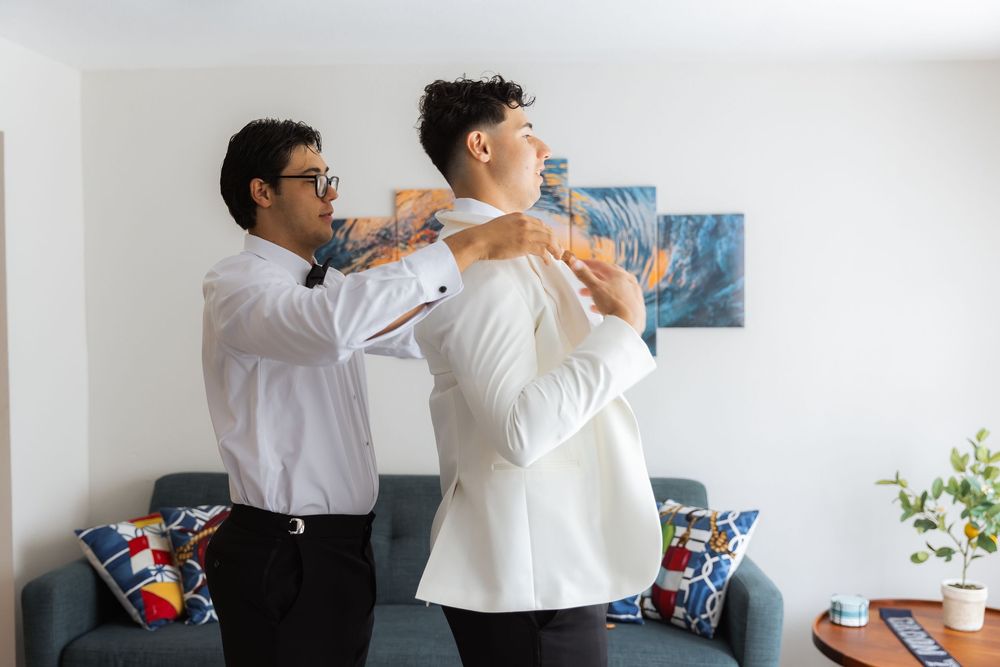 Two people in white dress shirts helping each other get ready in a living room with blue throw pillows on a couch.