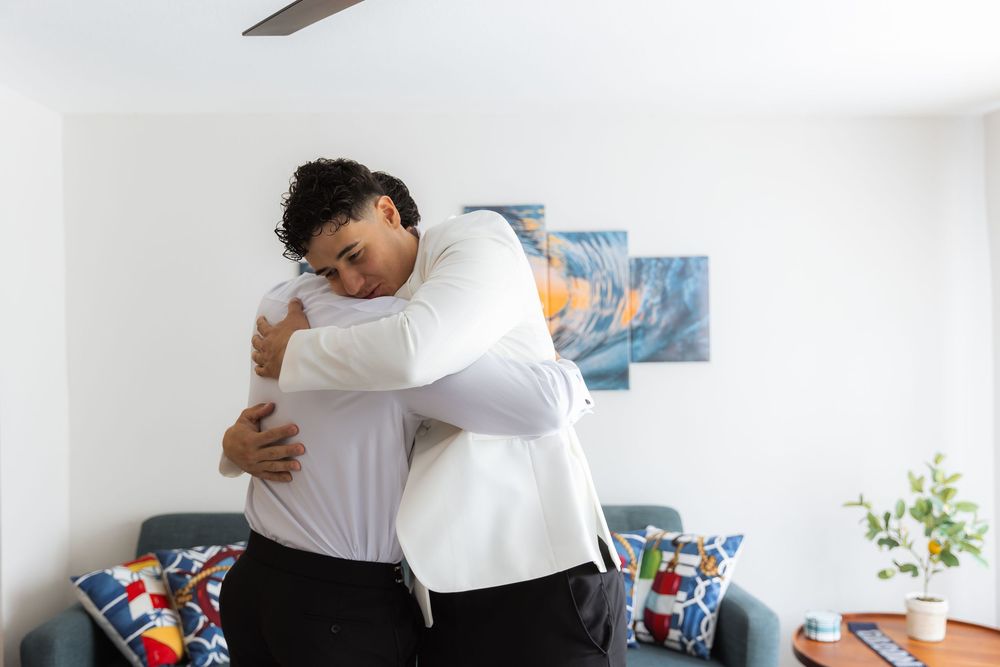Two people share a warm embrace while standing in front of a couch with colorful pillows in a bright room.
