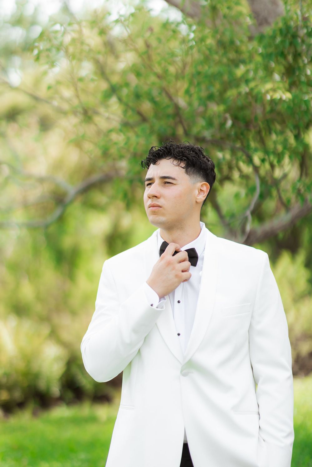 Figure in a crisp white tuxedo adjusts bowtie while standing in front of lush green garden backdrop.