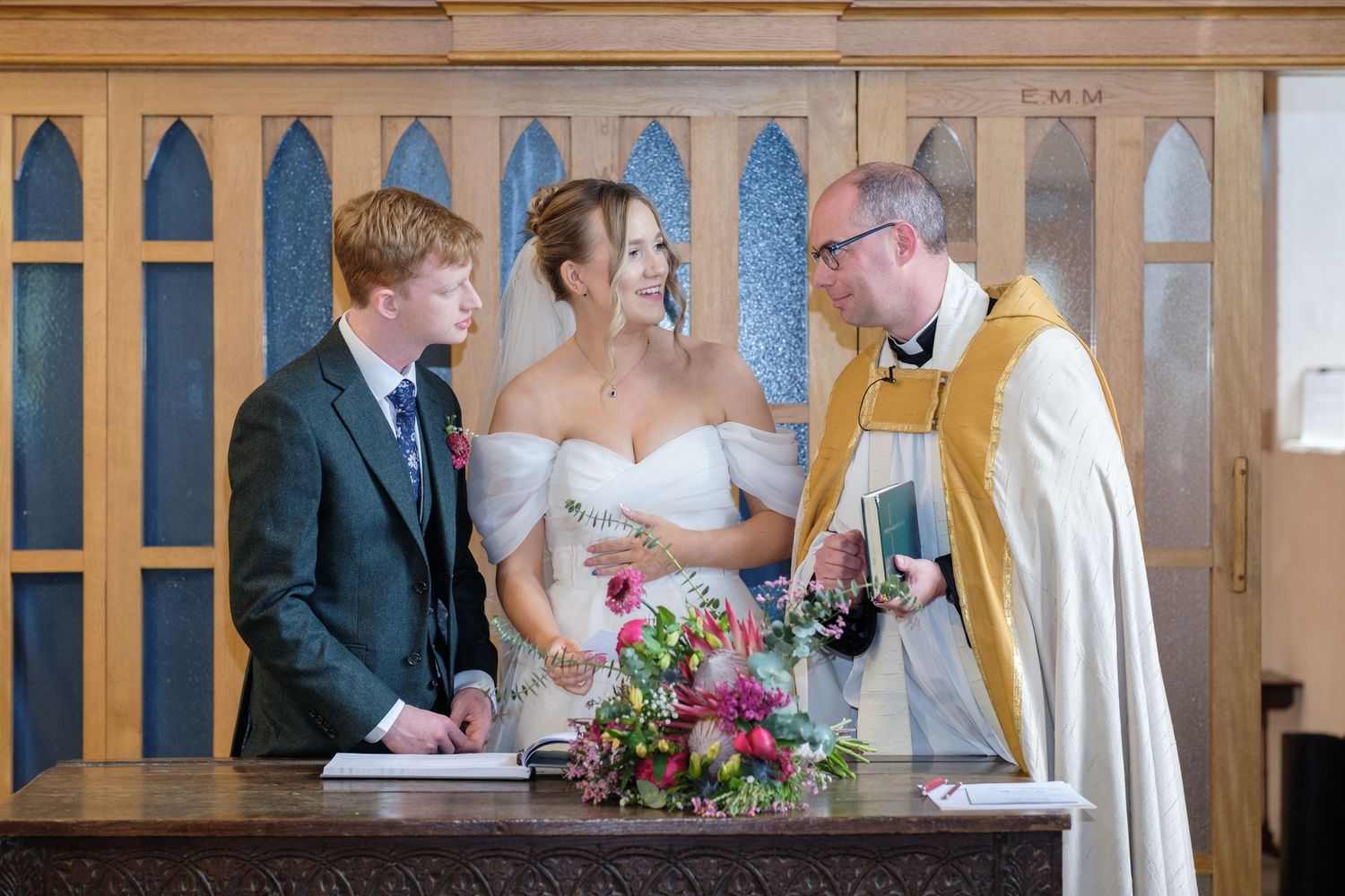 A couple renews their wedding vows during a ceremony with a priest in a church setting with wooden panels.