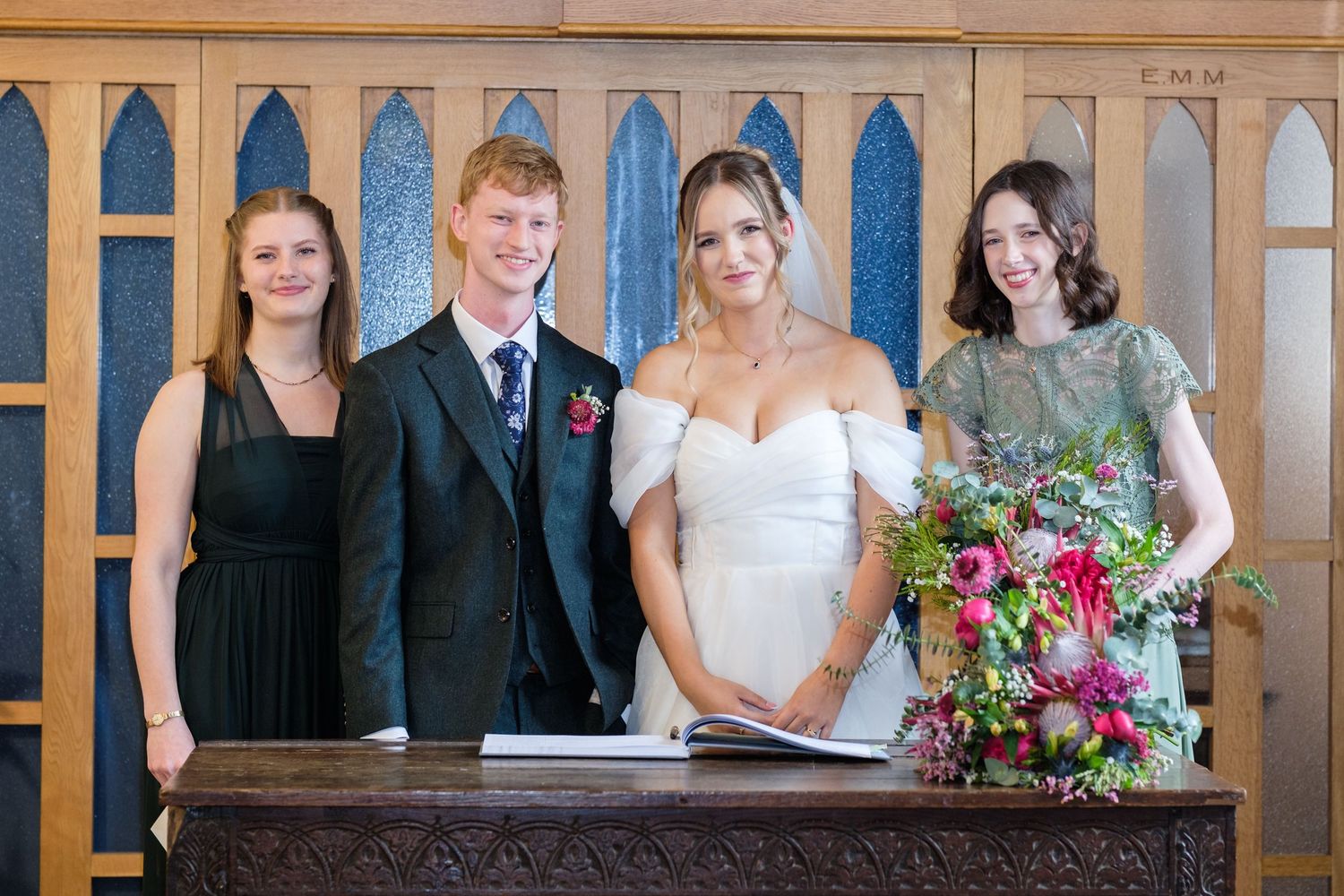 A wedding party stands together at a wooden altar in front of decorative arched windows during a ceremony.