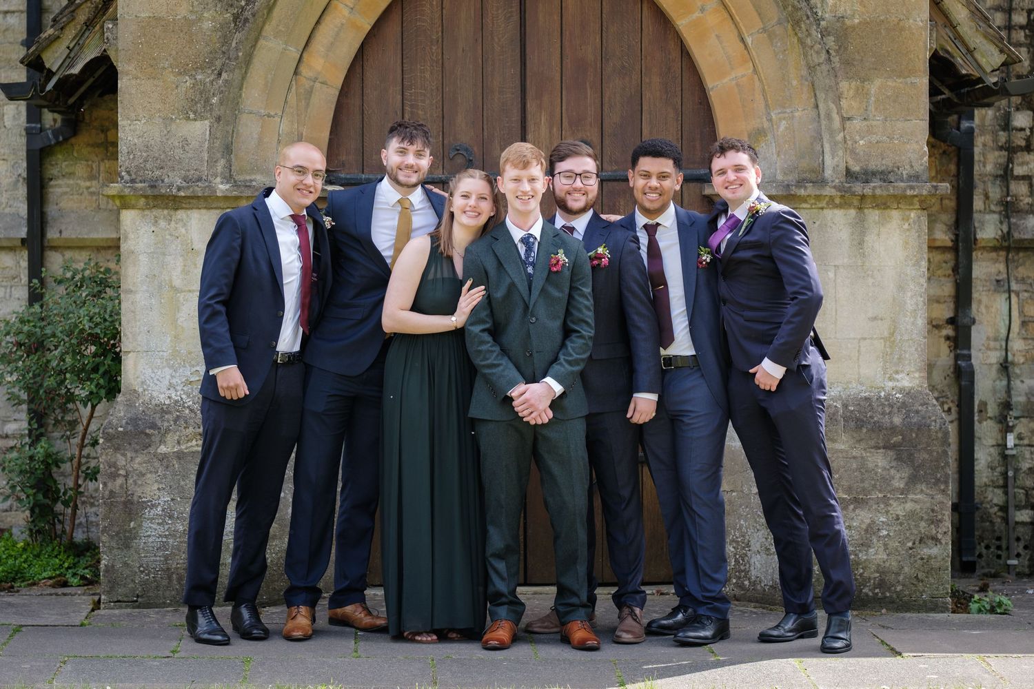 A group stands together formally posed in front of a stone archway wearing elegant suits and formal attire.