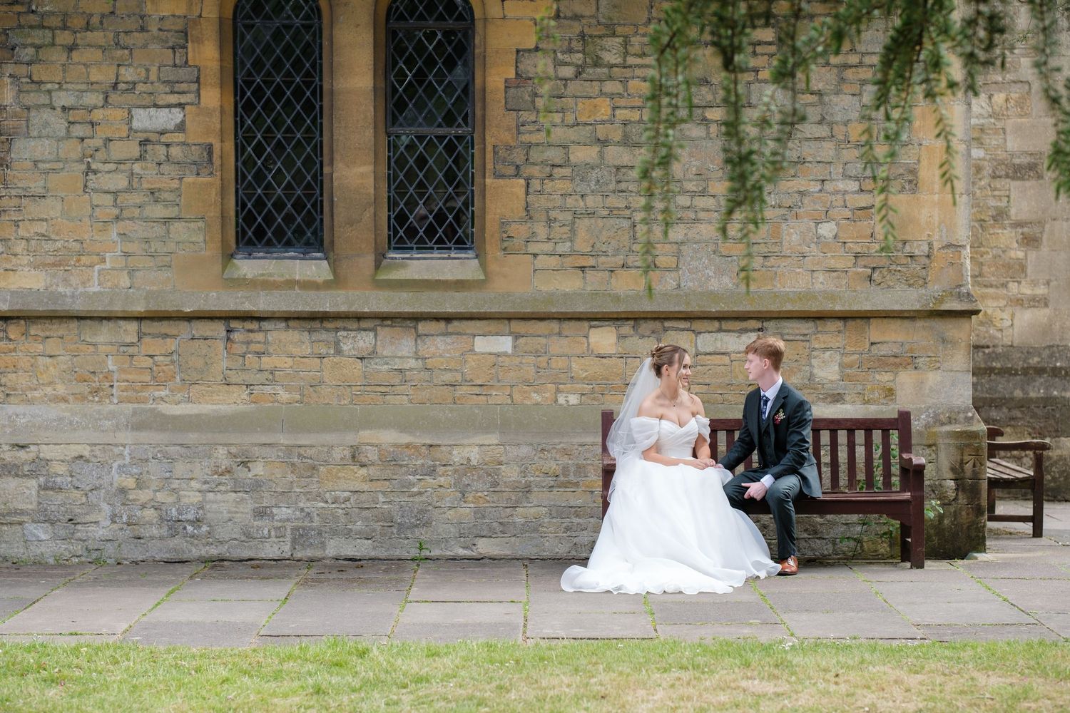 A couple sits together on a wooden bench outside a stone church building with gothic windows and climbing vines.