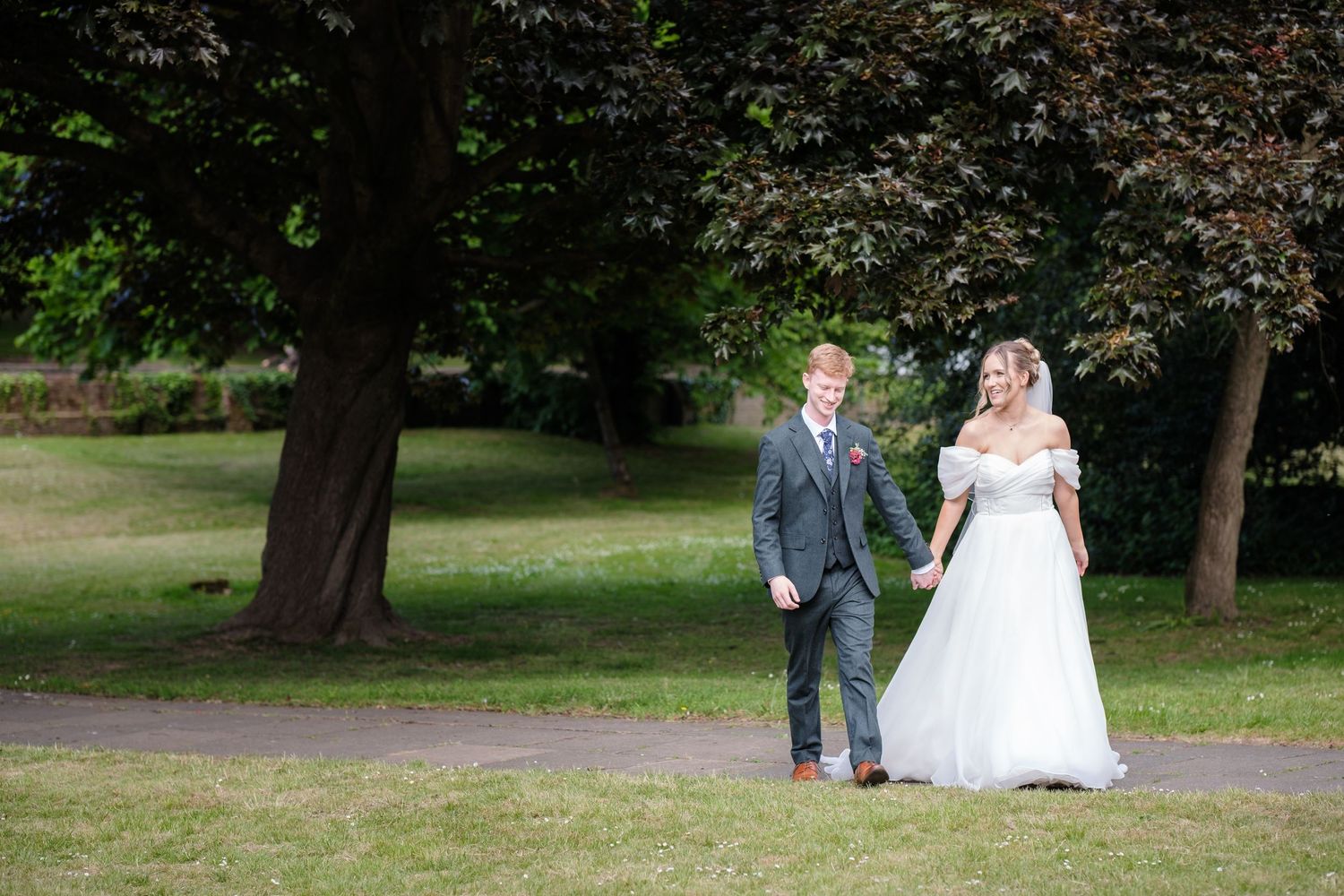 A newlywed couple walks hand in hand along a tree-lined park path on their wedding day.