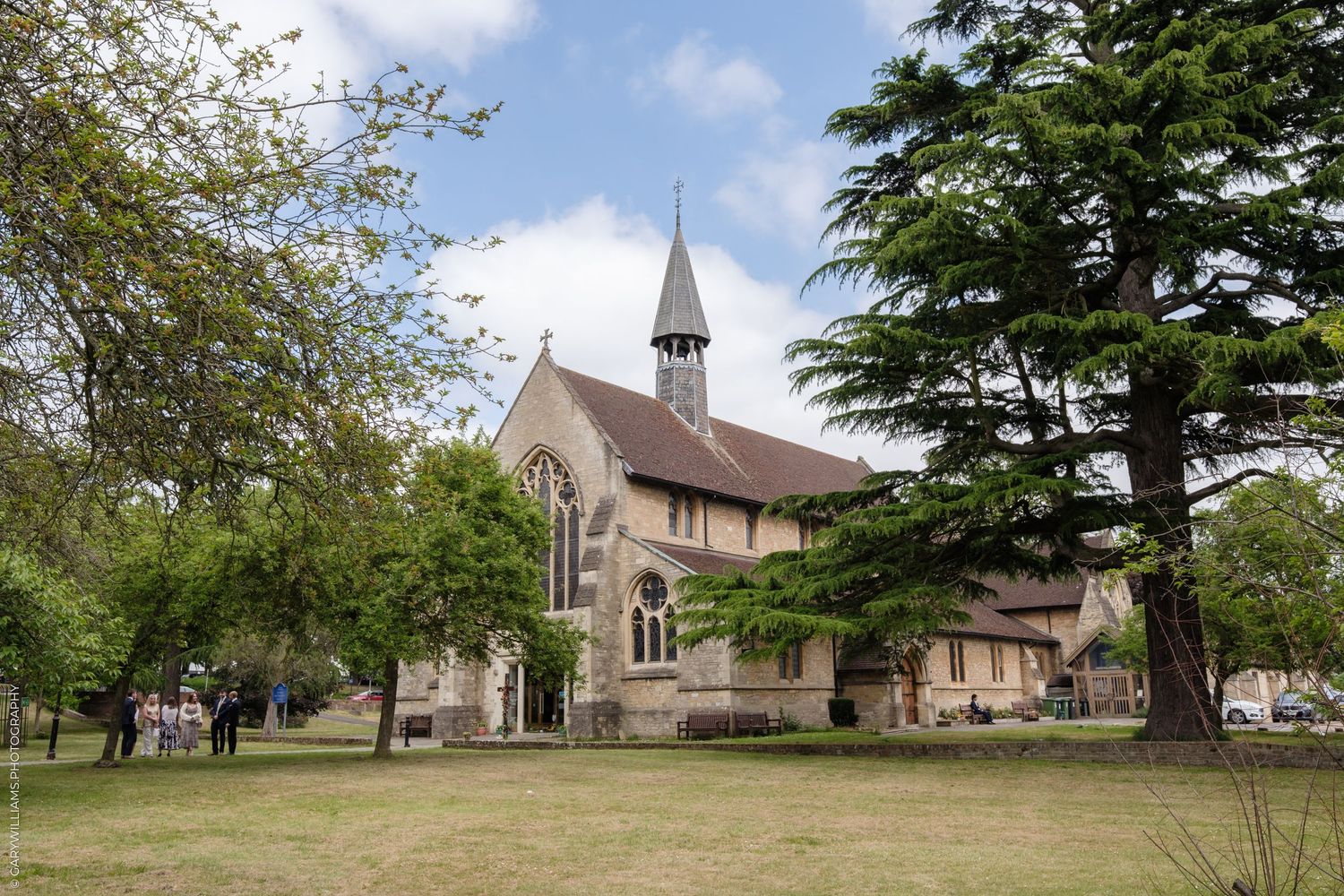 A tan stone church with a tall spire stands among green trees on a sunny day.