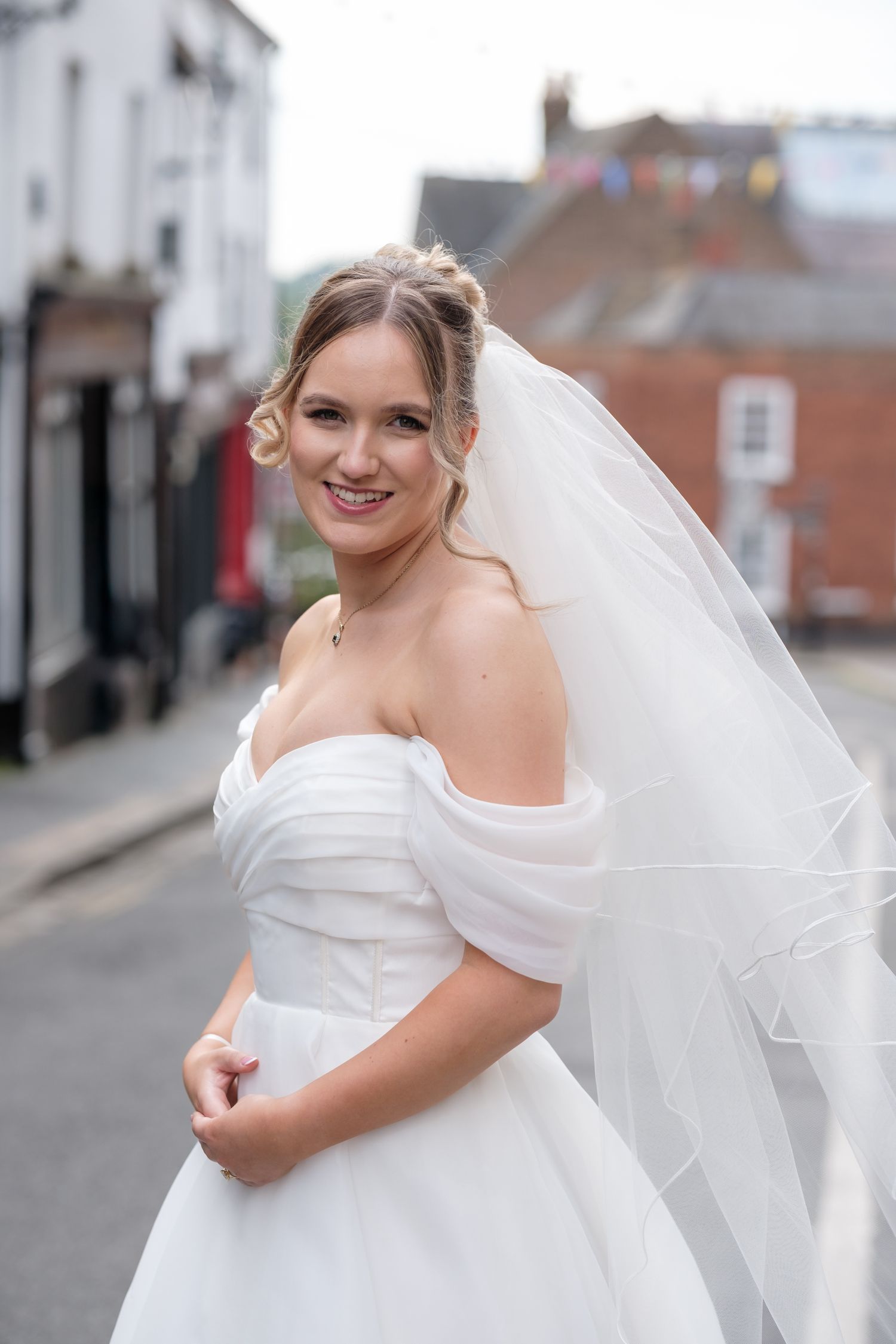 A bride wearing an elegant off-shoulder white wedding dress and flowing veil poses on a city street.