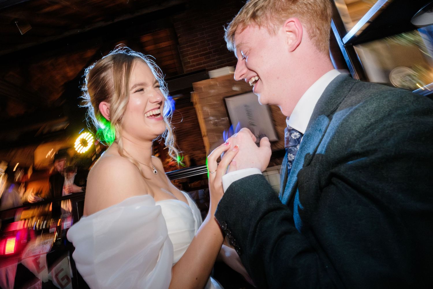Two people share a joyful dance moment together at a wedding reception with blurred lights in the background.