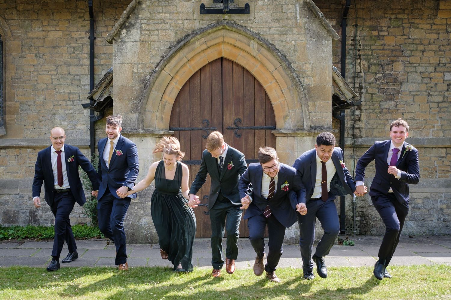 A wedding party jumps playfully outside a historic church with gothic architecture.