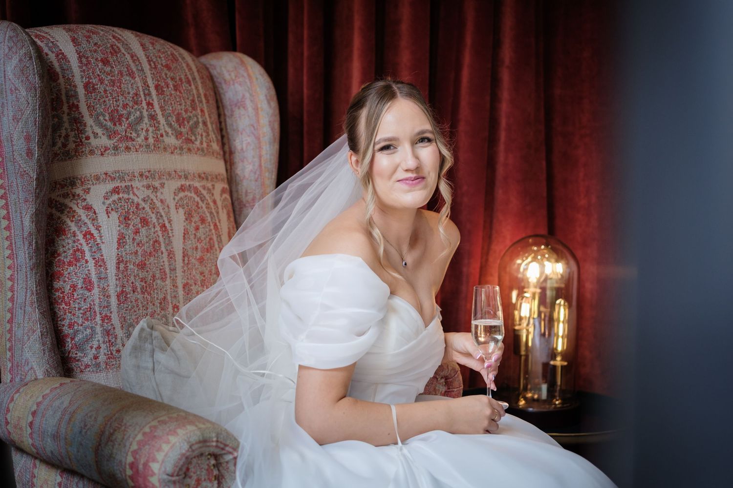 A bride in a white off-shoulder gown sits in an ornate chair holding a champagne glass next to a vintage lamp.