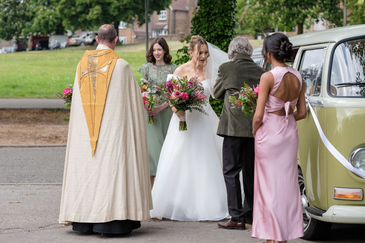 Wedding party gathers near vintage VW van with officiant in ceremonial robes and colorful bouquet.