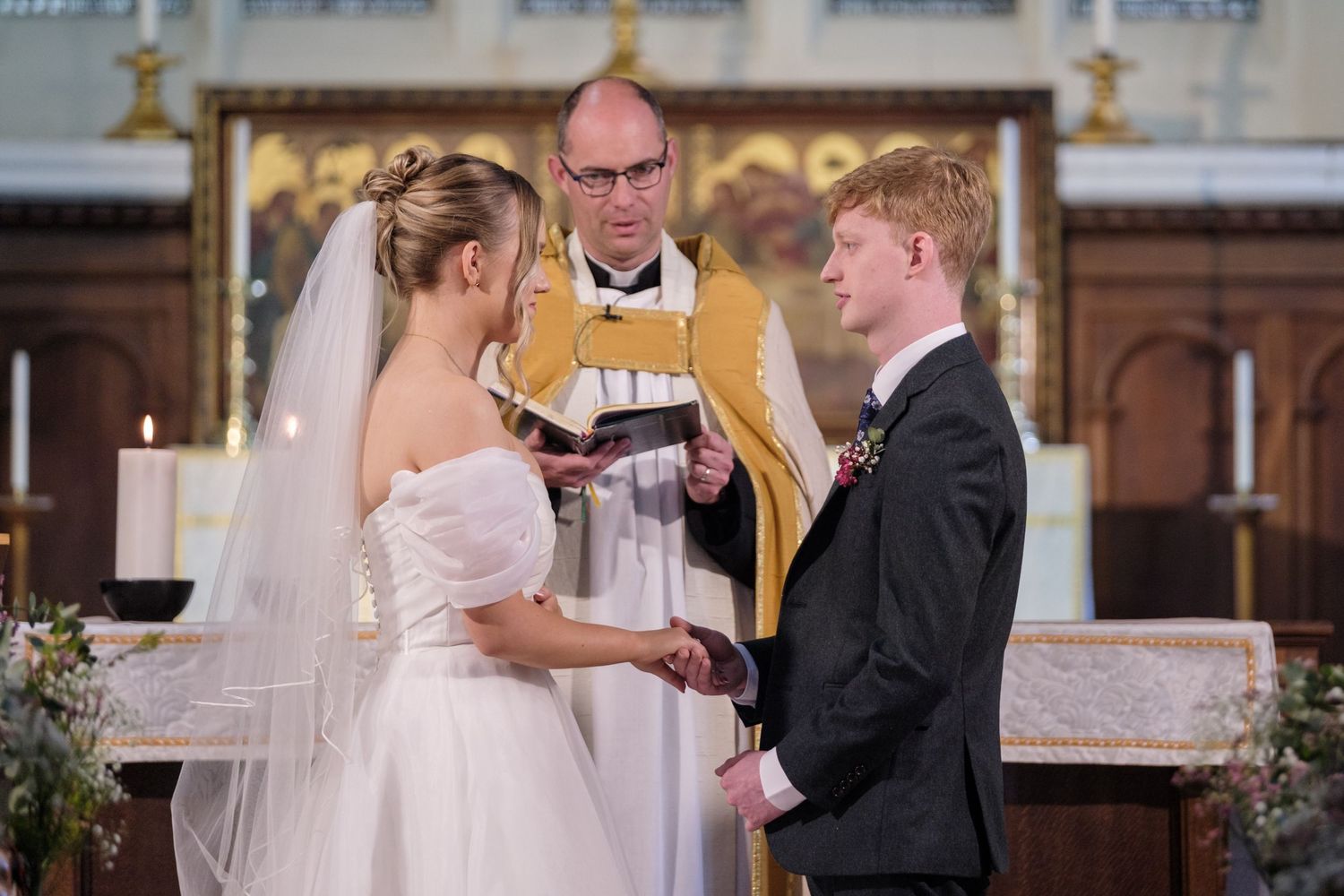 A wedding ceremony takes place in a traditional church with a minister in gold robes presiding over the service.