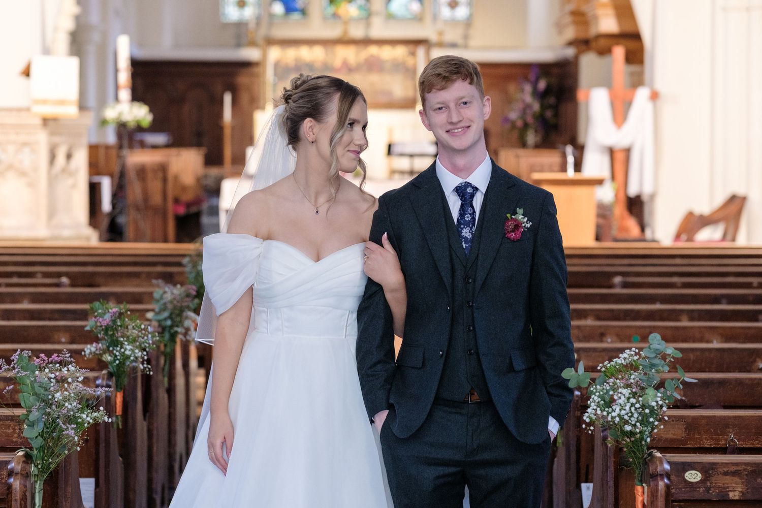 A couple stands together at a church altar, with the bride wearing an off-shoulder white gown and greenery decorations visible.