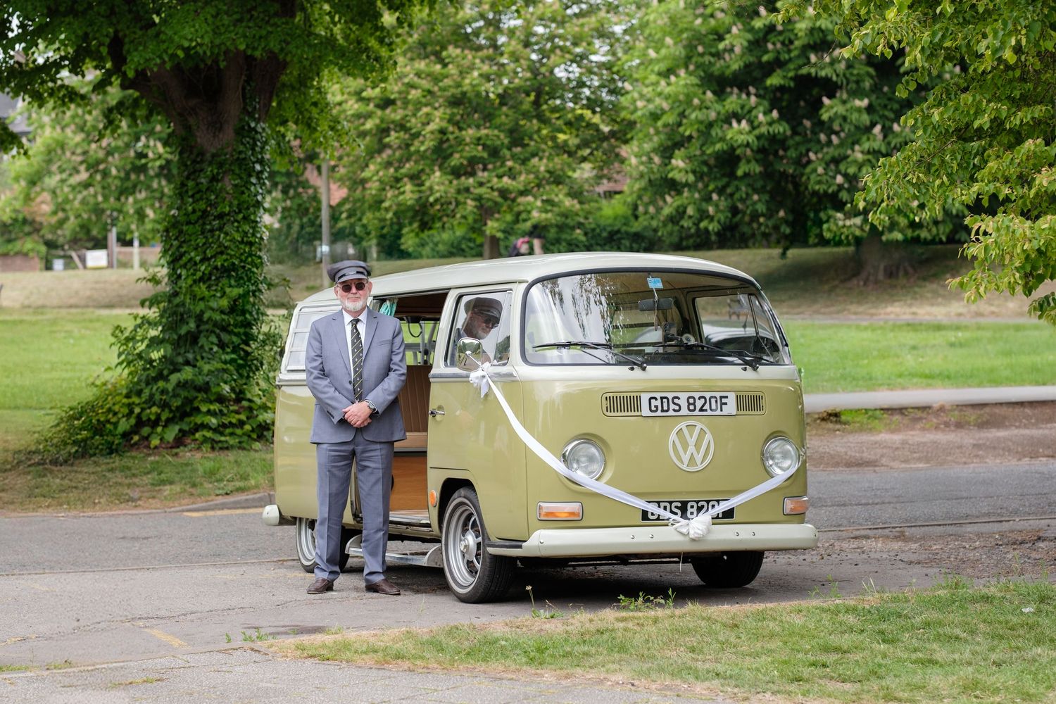 A vintage green and cream Volkswagen Type 2 bus parked on a tree-lined path in a park setting.
