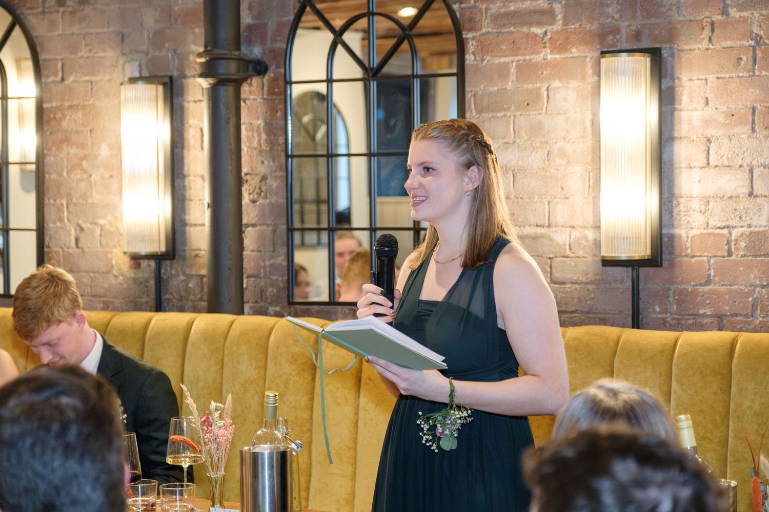 A server in a black dress takes an order at an elegant restaurant with exposed brick walls and sconce lighting.