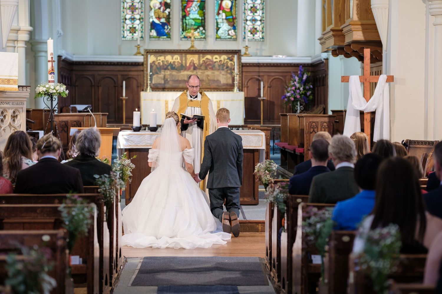 Traditional wedding ceremony inside a historic church with stained glass windows and floral decorations along the aisle.