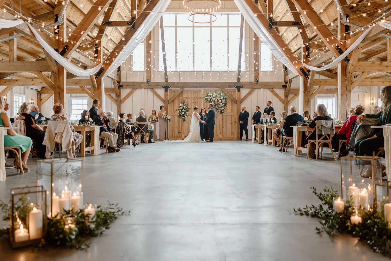 Rustic barn wedding ceremony setup with wooden beams, lantern aisle decor and string lights overhead.