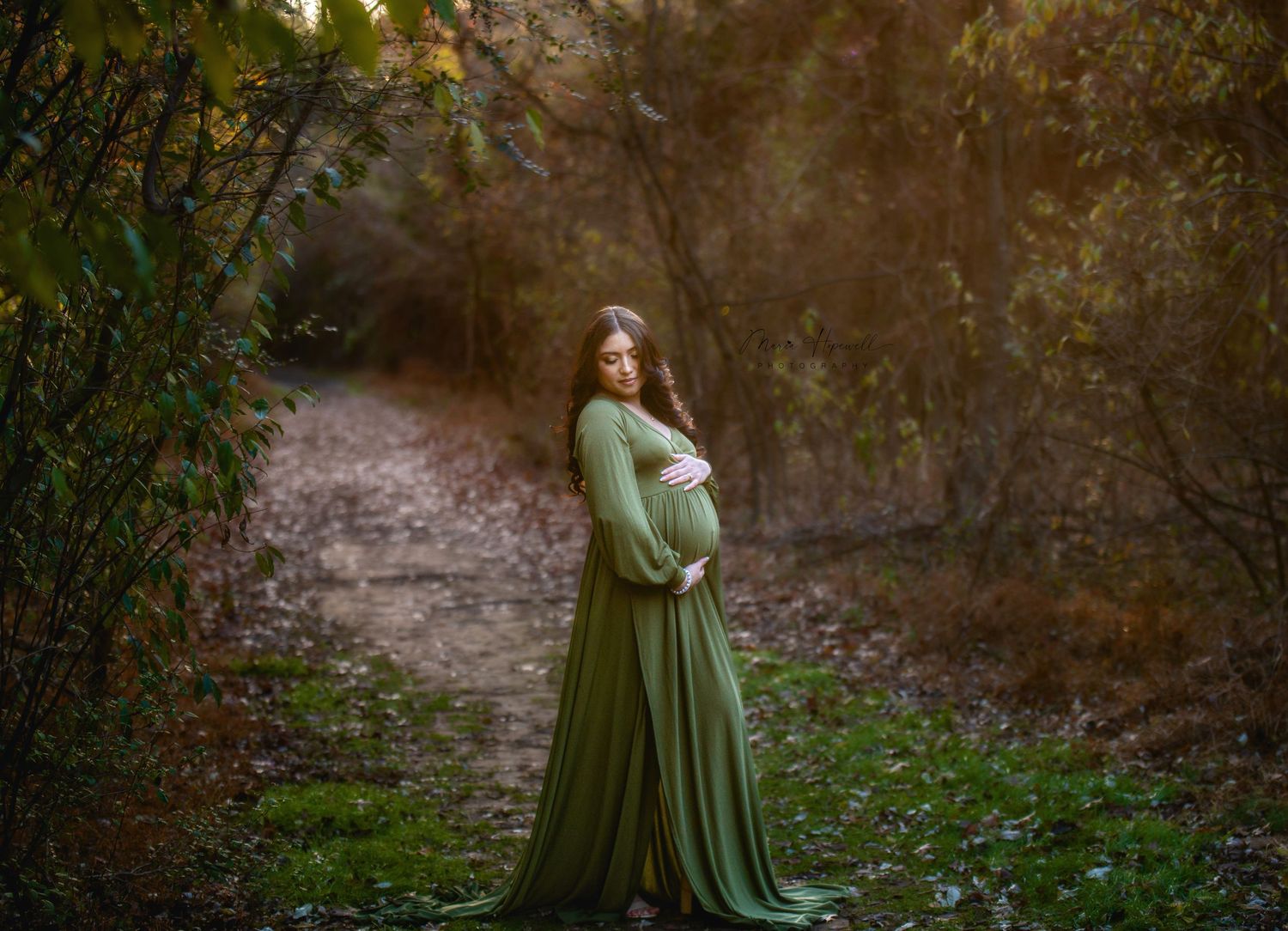 A maternity portrait in a flowing sage green dress on a wooded forest path.