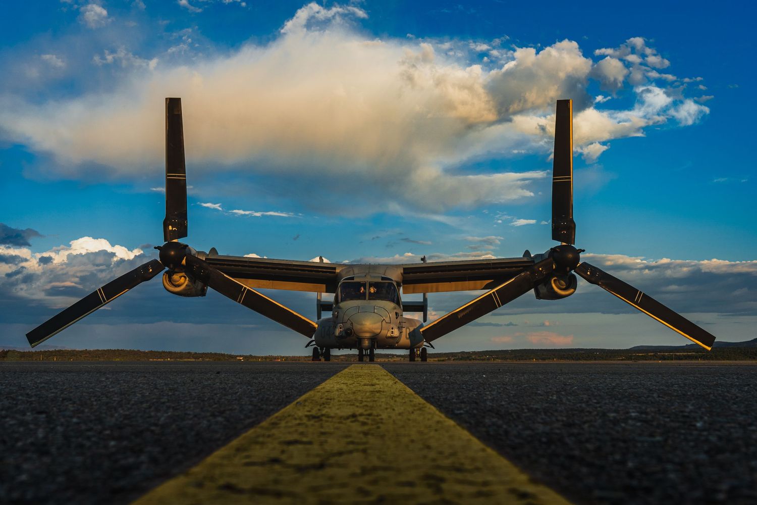 Military V-22 Osprey aircraft with dual rotors sits on runway under dramatic blue sky with puffy white clouds.