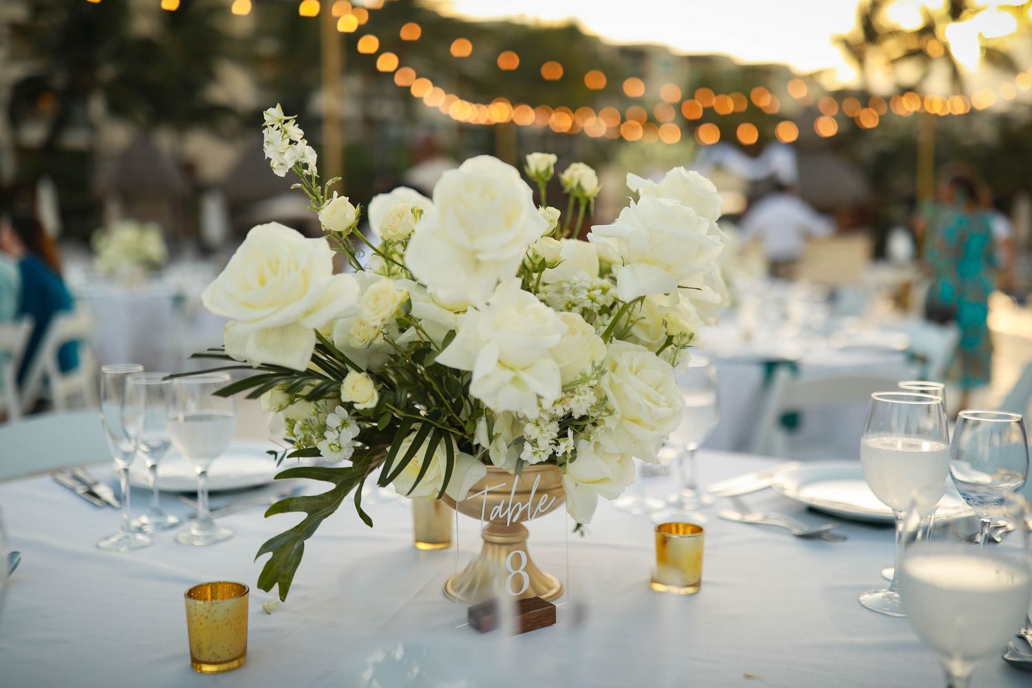 Wedding reception table decorated with white floral centerpieces and string lights overhead.