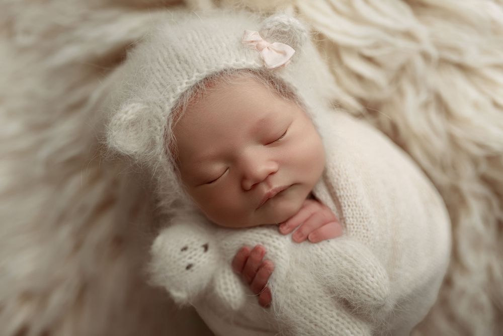 Sleeping newborn baby in white knit bear bonnet and wrap, cradling a tiny teddy bear on a fluffy cream fur backdrop.