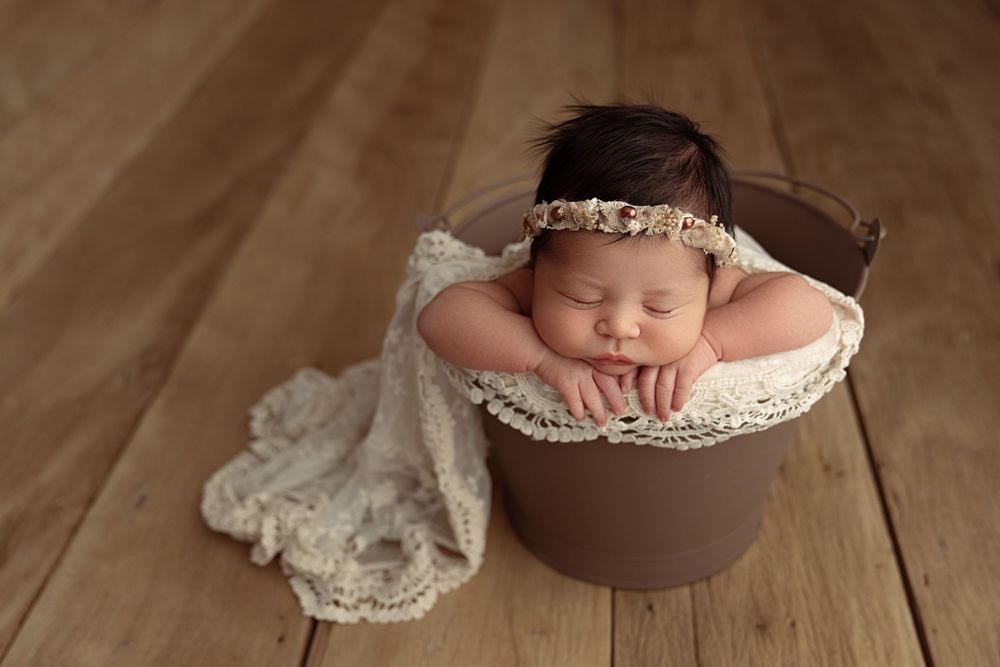 A sleeping baby nestled in a brown wooden bowl with delicate lace fabric on a wooden floor. – newborn photographer Paloma Schell Houston TX