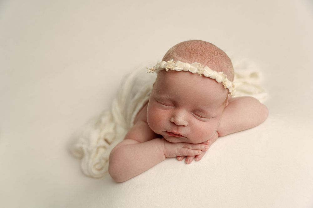 A sleeping newborn baby wearing a delicate cream headband rests peacefully on soft white fabric– newborn photographer Paloma Schell Houston