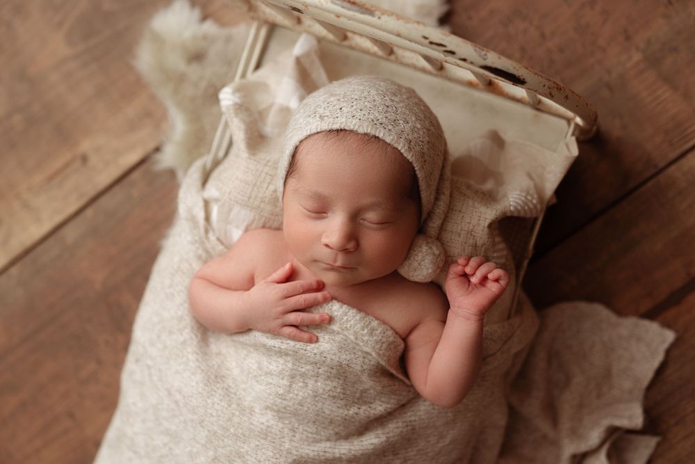 A sleeping newborn wrapped in soft white fabric lies on a textured wooden surface. – newborn photographer Paloma Schell Houston TX