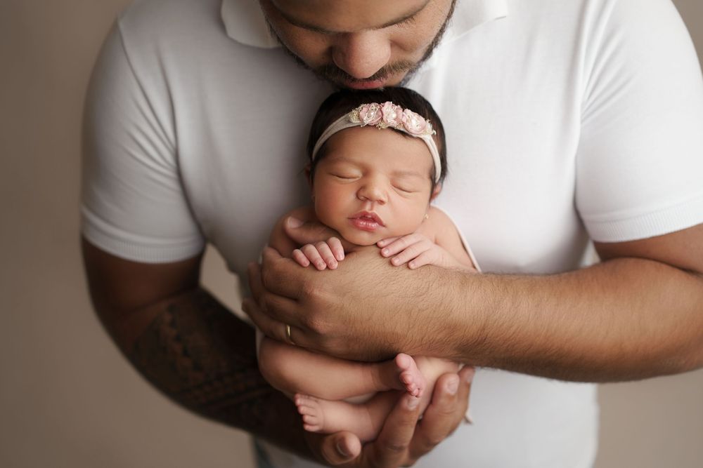 A sleeping newborn baby in white is held in protective arms against a neutral background. – newborn photographer Paloma Schell Houston TX