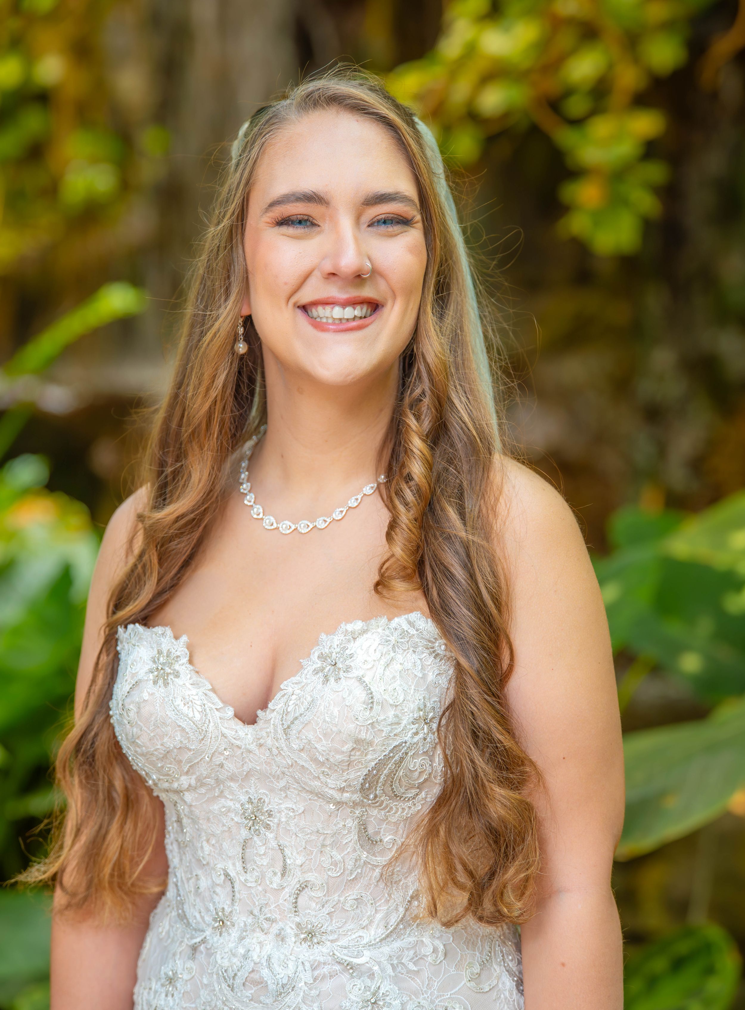 A bride in a white lace wedding dress poses outdoors with natural greenery in the background.
