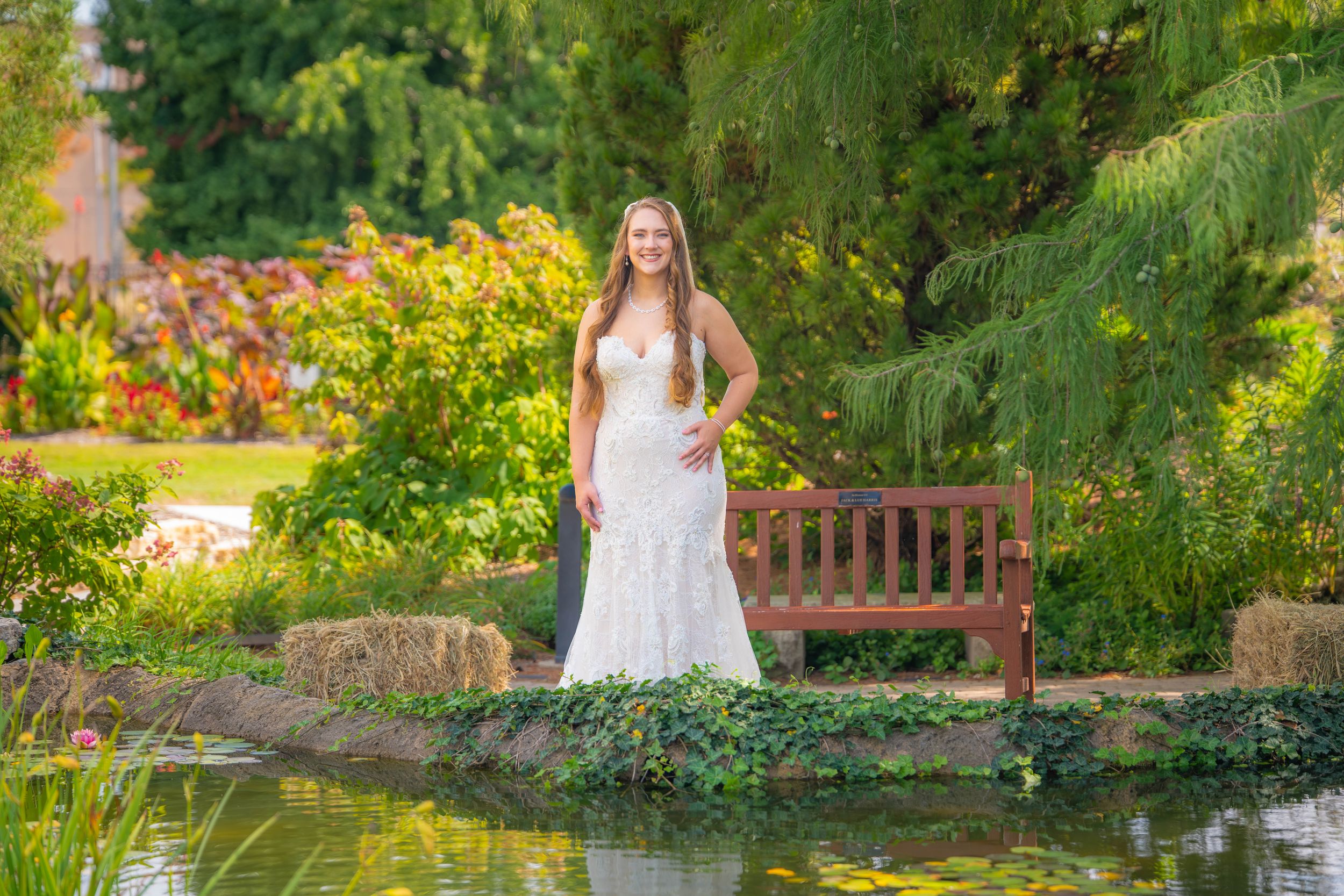 A tranquil garden scene with a wooden bench beside a pond surrounded by lush greenery and blooming flowers.