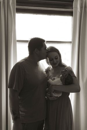 Family members stand by window holding newborn baby in backlit indoor setting.