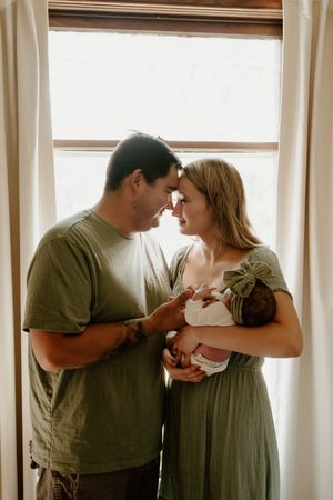 Couple in casual attire shares intimate moment with newborn by bright window.