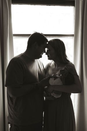Silhouetted figures stand together by window sharing tender moments with infant.