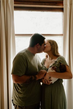 Parents share loving kiss while holding newborn baby near sunlit window.