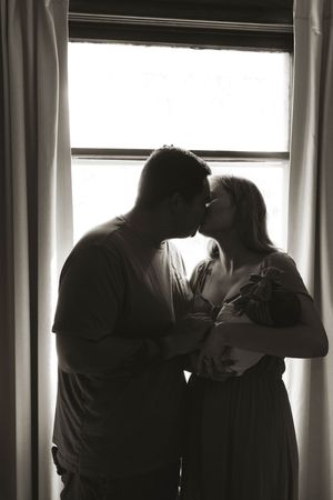 Silhouetted family members share tender moments with baby by window.