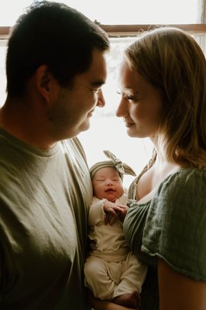 Parents share intimate moment with newborn baby in natural window light.