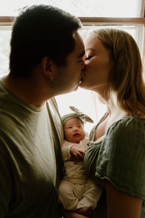 Intimate family moment by a sunlit window as parents share tender kisses while holding their newborn baby.