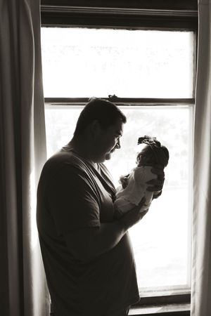 Silhouetted figures photograph newborn baby by bright window.