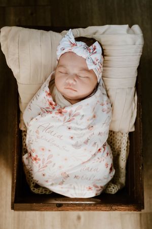 Peaceful newborn sleeping in wooden crate wrapped in delicate floral swaddle.