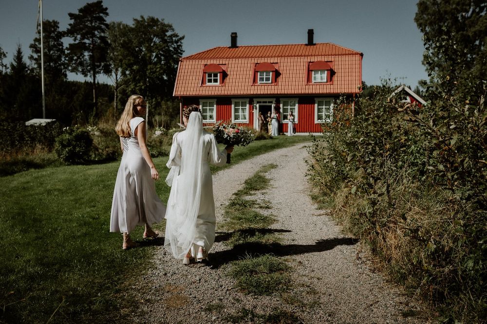 Brud med slöja går med en av sina bröllopsgäster mot ett rött hus på Näs Herrgård, Mullsjö.