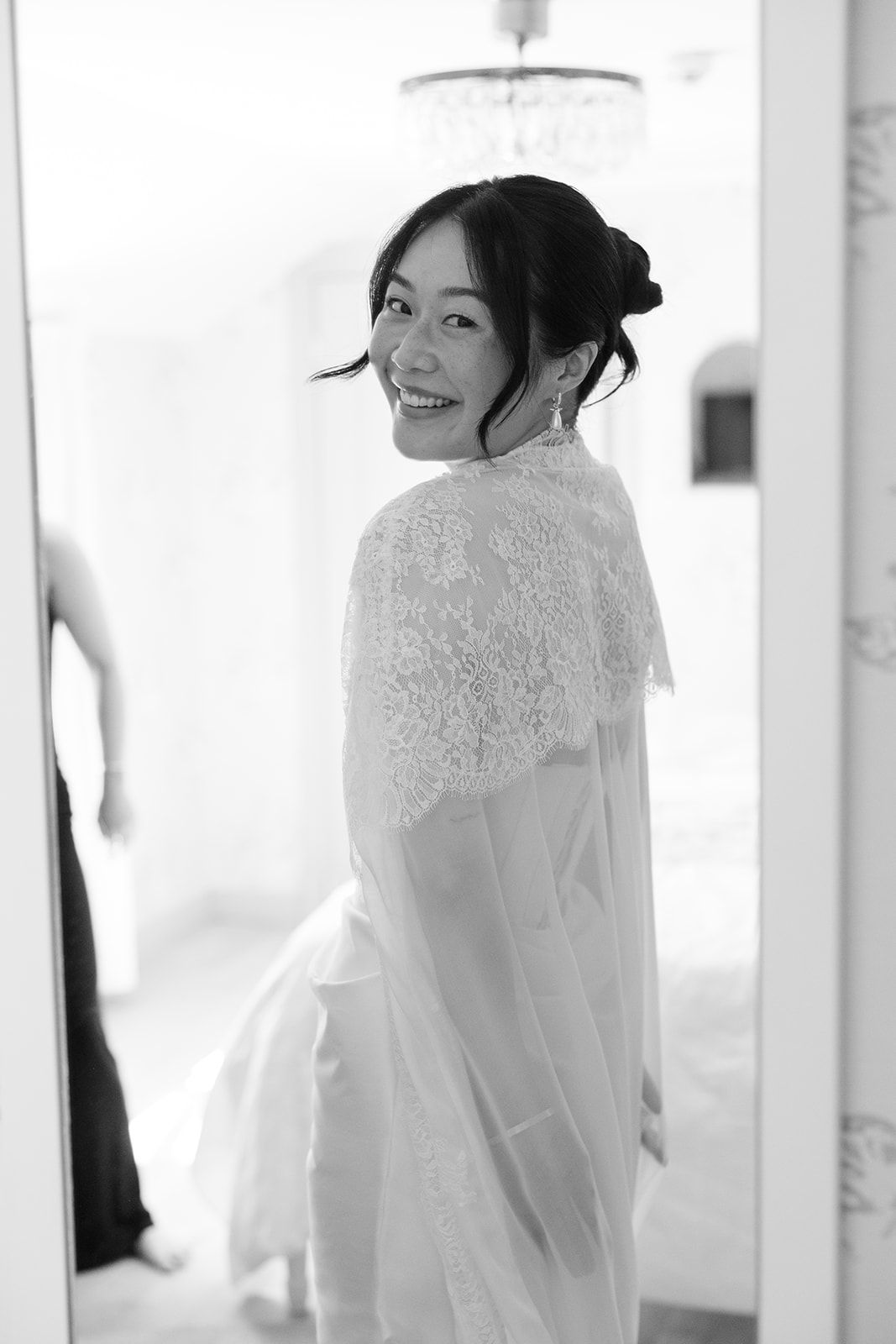 A bride in a delicate white lace wedding dress smiles softly in a bright, naturally lit room.