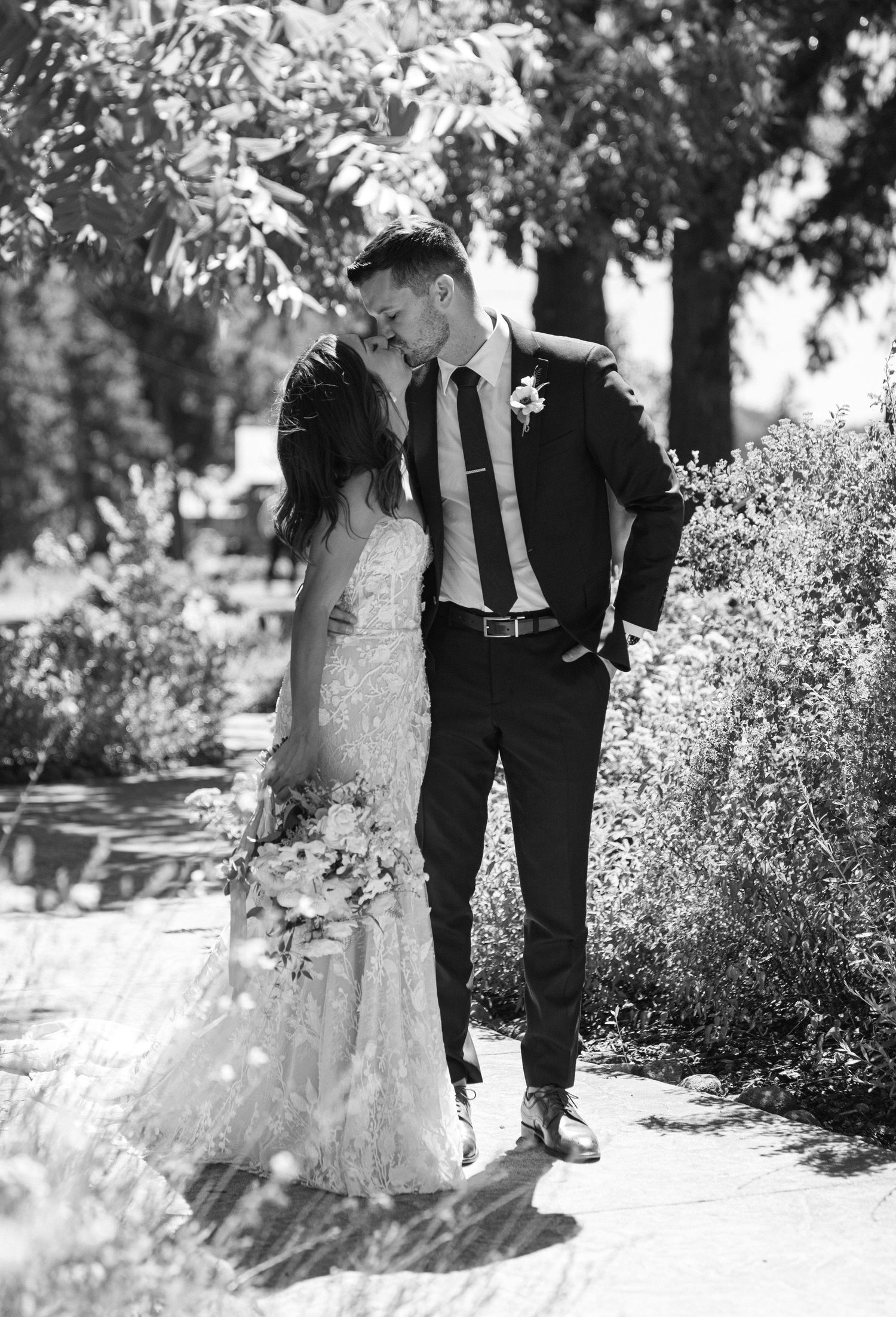 A couple in wedding attire strolls together through a garden path in black and white photography.