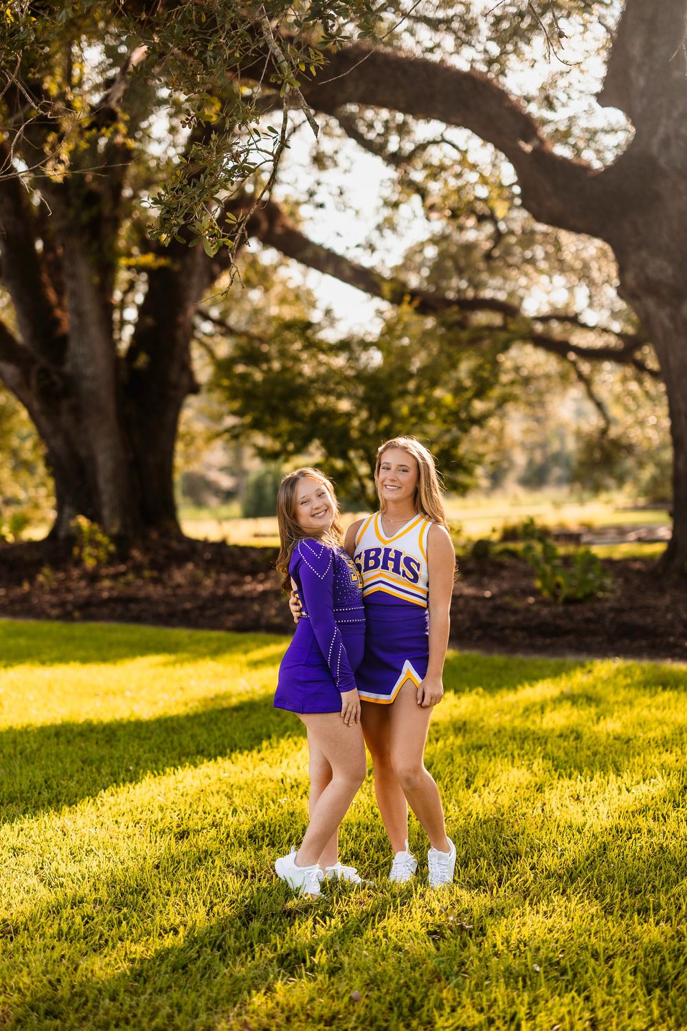 Two cheerleaders in blue uniforms sit together on grassy hill during golden hour.