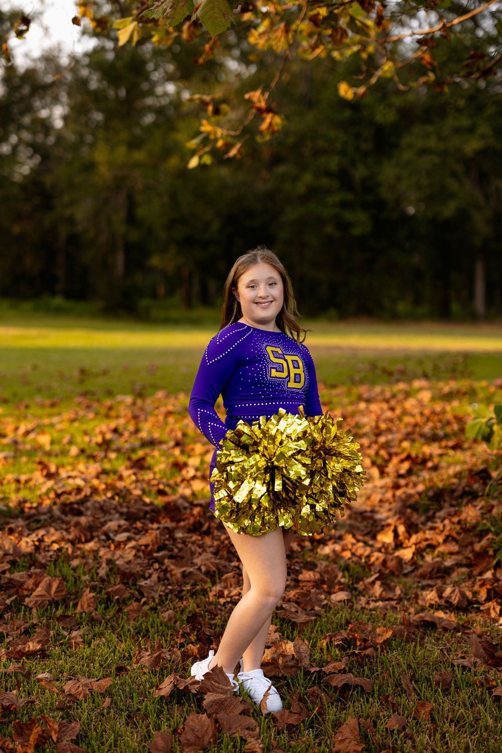 Cheerleader in purple uniform poses with gold pom poms among autumn leaves in outdoor setting.