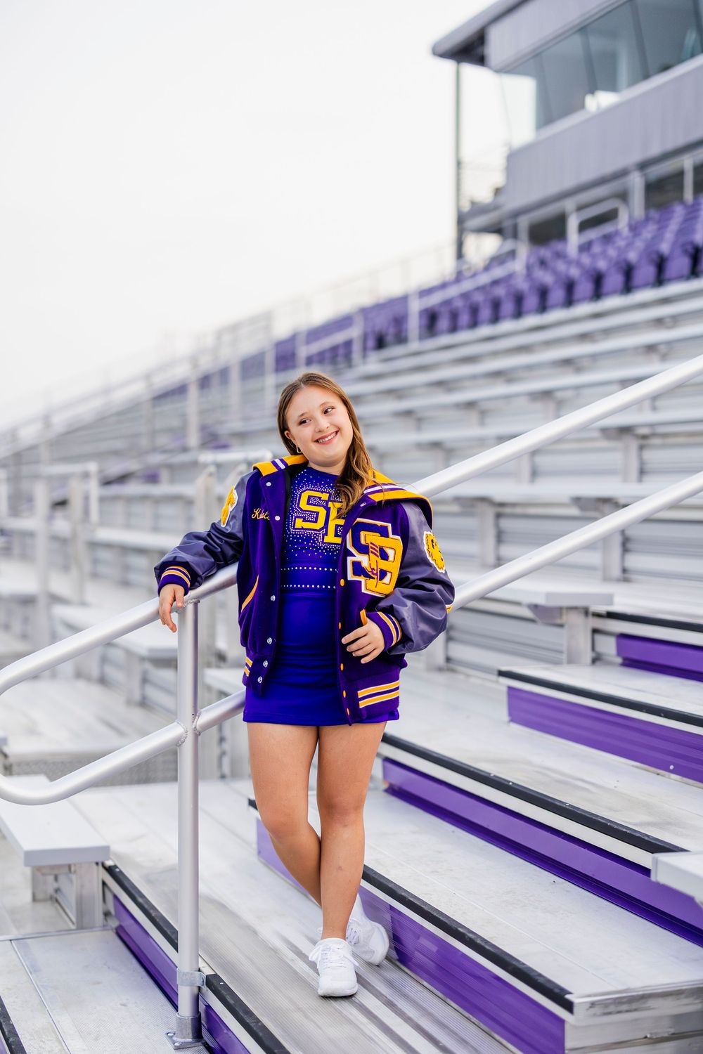 Cheerleader in letterman jacket poses on purple and white stadium bleachers.