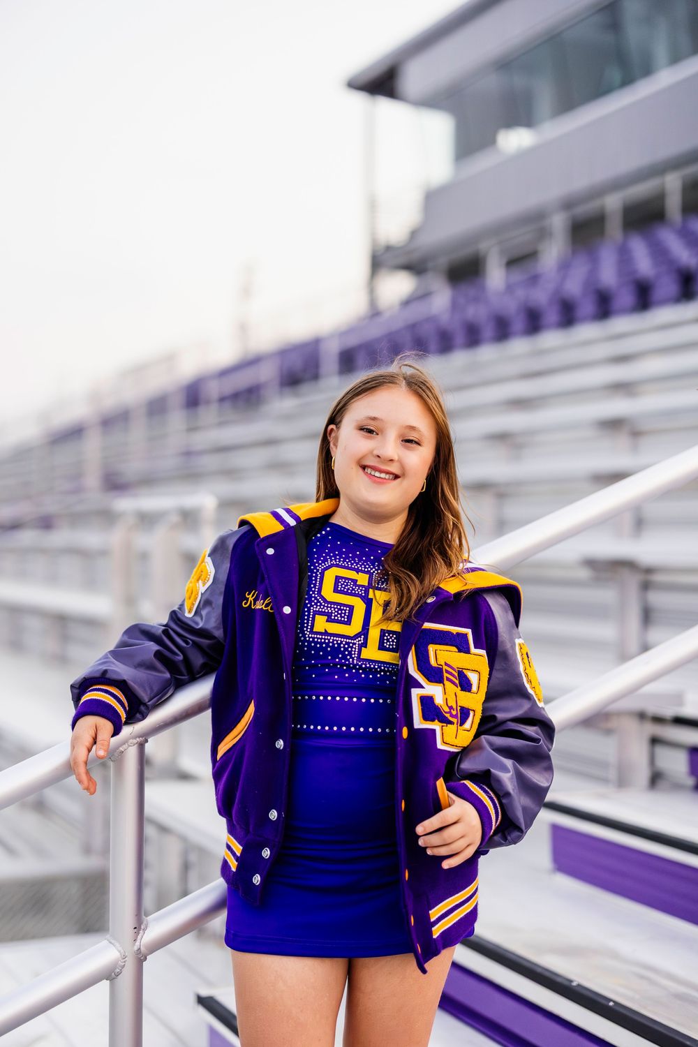 Cheerleader in navy varsity jacket leans against stadium railing.