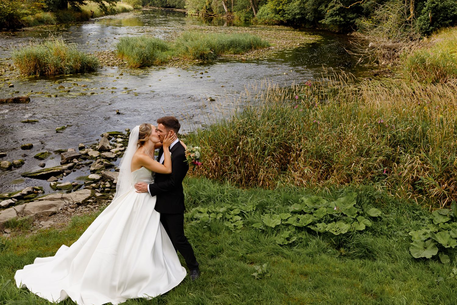 Couple in formal wedding attire embraces on a grassy riverbank surrounded by natural scenery.